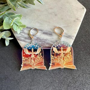 May include: A pair of iridescent earrings featuring a dragon emerging from an open book with a crescent moon design. The earrings are made of clear acrylic and have silver hooks.