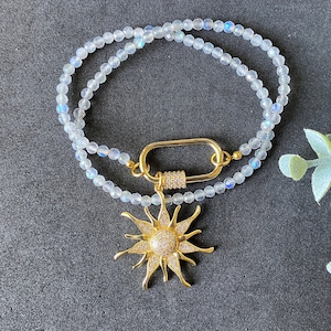 以下が含まれることがあります： 小さな虹色のビーズが2連になった繊細なネックレス。ゴールドカラーの留め金で連結され、輝くクリスタルで飾られたサンバーストペンダントが付いています。太陽のペンダントには、中央にクリスタルのクラスターがあります。