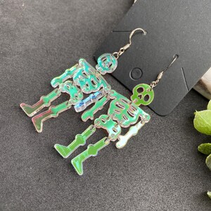 May include: A pair of dangling earrings featuring articulated skeleton figures made from iridescent, semi-transparent acrylic material. Each skeleton has a distinct skull, rib cage, and leg segments connected by small silver-toned metal jump rings.