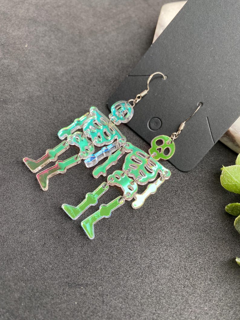 May include: A pair of dangling earrings featuring articulated skeleton figures made from iridescent, semi-transparent acrylic material. Each skeleton has a distinct skull, rib cage, and leg segments connected by small silver-toned metal jump rings.