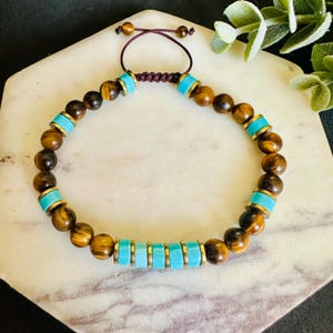 May include: A beaded bracelet featuring round brown tiger eye stones and cylindrical turquoise-colored beads separated by thin gold-tone metal spacers. The bracelet is secured with a dark brown adjustable cord closure with two small beads at the ends.