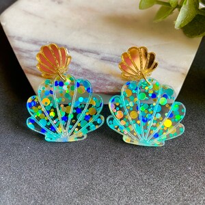 May include: A pair of iridescent blue and green seashell earrings with gold accents. The seashells are filled with iridescent glitter and have a clear, translucent finish.