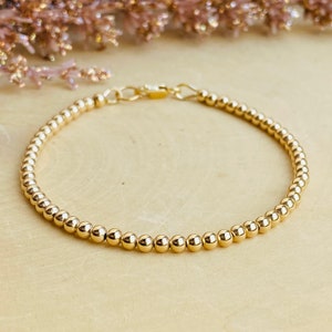 May include: A gold beaded bracelet with a clasp closure. The bracelet is made of small, round beads that are evenly spaced.