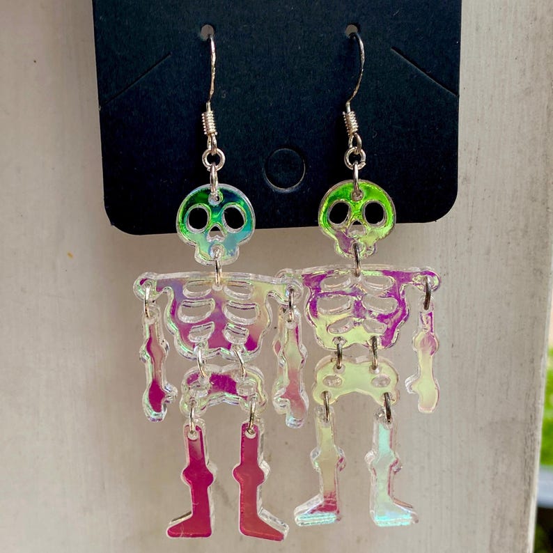 May include: A pair of dangling earrings featuring iridescent, holographic skeleton figures. Each skeleton is constructed from multiple articulated pieces connected by silver-toned metal jump rings, suspended from standard silver-toned earring hooks.