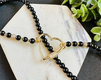 Op de afbeelding: Een zwarte kralenketting met een gouden hartvormige sluiting. De sluiting is versierd met kleine, heldere stenen.