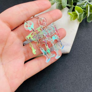 May include: Dangle earrings featuring articulated skeleton figures made of iridescent, semi-transparent acrylic material. Each skeleton consists of a skull, ribcage, and leg segments connected by silver-toned metal jump rings for movement.