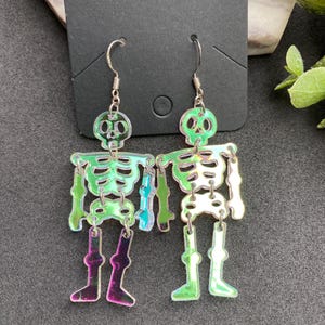 May include: A pair of dangling earrings featuring articulated skeleton figures made from iridescent, semi-transparent acrylic material. The skeletons have a reflective, color-shifting surface that displays green, purple, and silver tones, suspended from silver-toned metal fishhook ear wires.