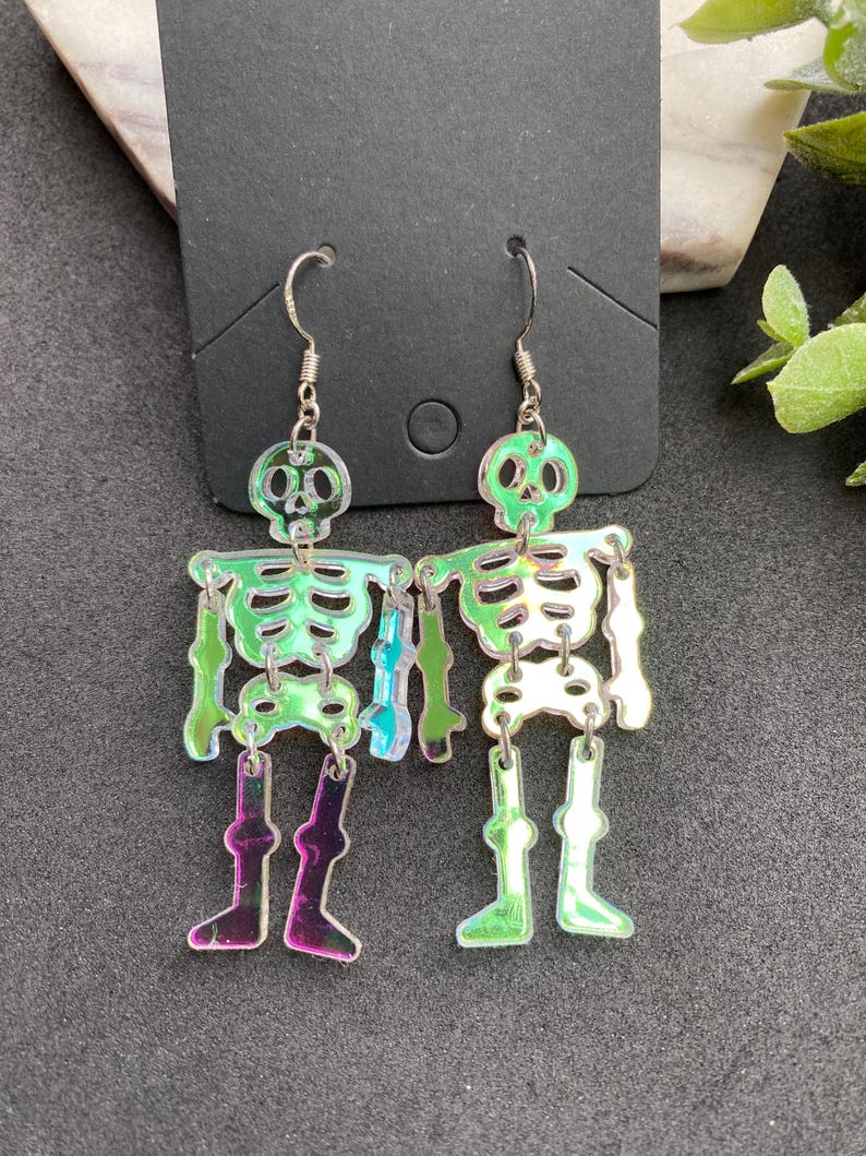 May include: A pair of dangling earrings featuring articulated skeleton figures made from iridescent, semi-transparent acrylic material. The skeletons have a reflective, color-shifting surface that displays green, purple, and silver tones, suspended from silver-toned metal fishhook ear wires.
