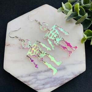 May include: A pair of dangle earrings featuring iridescent, semi-transparent skeleton figures. Each skeleton is constructed from articulated segments connected by silver-toned metal jump rings, suspended from simple silver-toned metal earring hooks.
