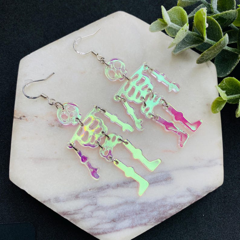 May include: A pair of dangle earrings featuring iridescent, semi-transparent skeleton figures. Each skeleton is constructed from articulated segments connected by silver-toned metal jump rings, suspended from simple silver-toned metal earring hooks.
