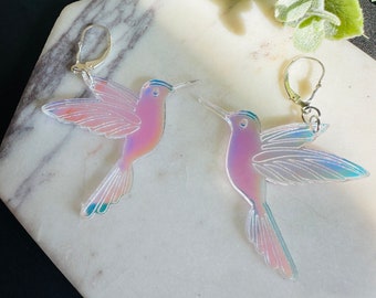 May include: A pair of iridescent hummingbird earrings with silver hooks. The hummingbirds are made of clear acrylic with a rainbow iridescent finish.