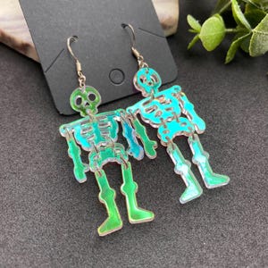May include: A pair of iridescent green skeleton earrings. The earrings are made of acrylic and have a silver hook.