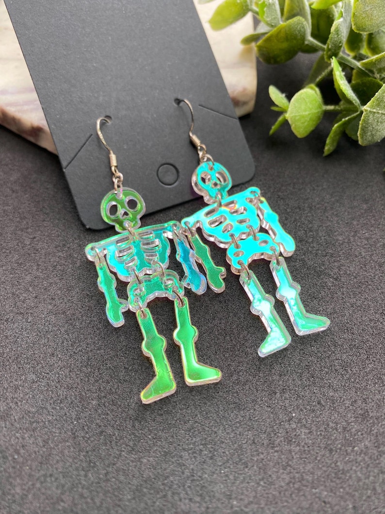 May include: A pair of dangling earrings featuring iridescent skeleton figures with a green tint. The skeletons are constructed from multiple articulated acrylic pieces connected by small metal jump rings, suspended from silver-toned metal earring hooks.