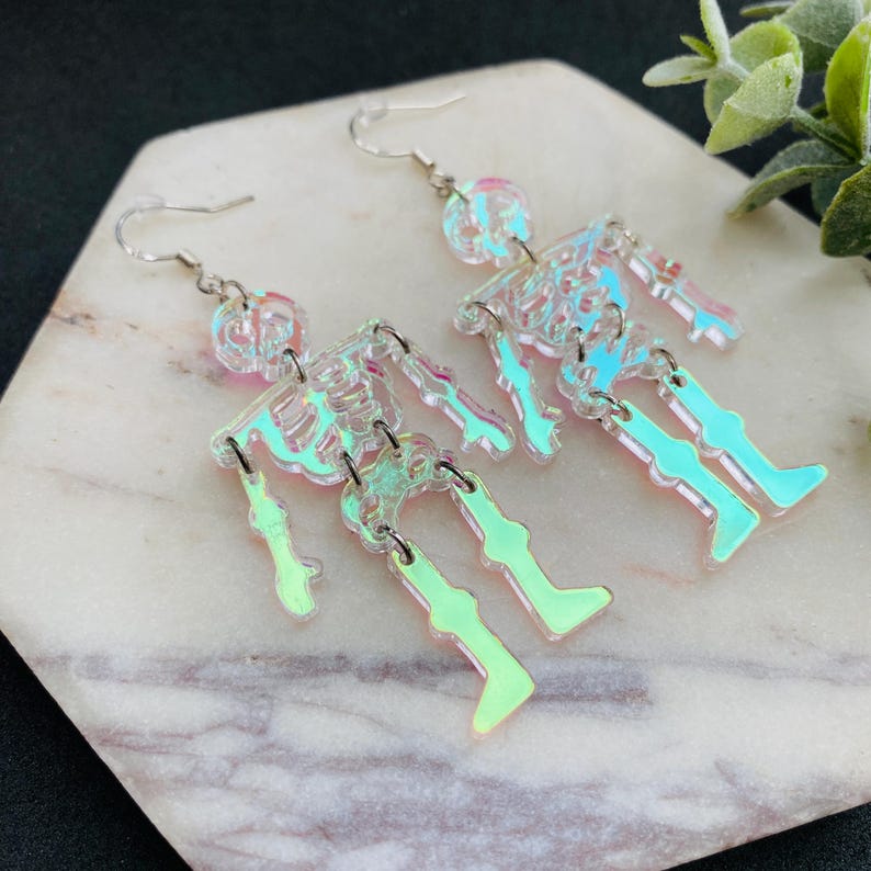 May include: A pair of dangling earrings featuring articulated skeleton figures made from iridescent, semi-transparent material. Each skeleton has a skull, torso, and leg segments connected by silver-toned metal jump rings and silver-toned ear hooks.