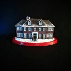 May include: A miniature replica of a two-story house with a red roof, white trim, and snow-covered ground. The house has a front porch with a wreath on the door and a red base.