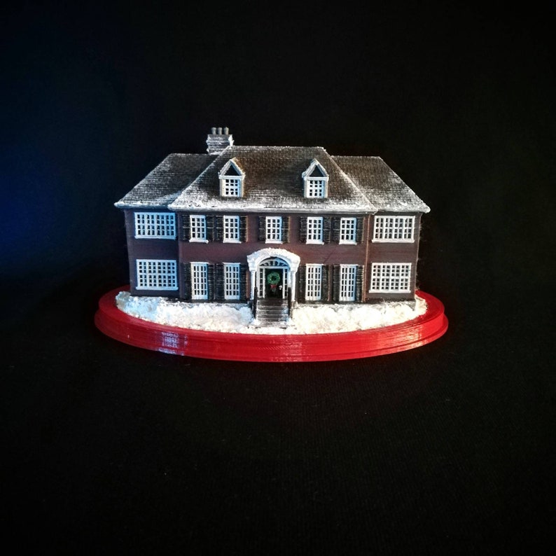 May include: A miniature replica of a two-story house with a red roof, white trim, and snow-covered ground. The house has a front porch with a wreath on the door and a red base.