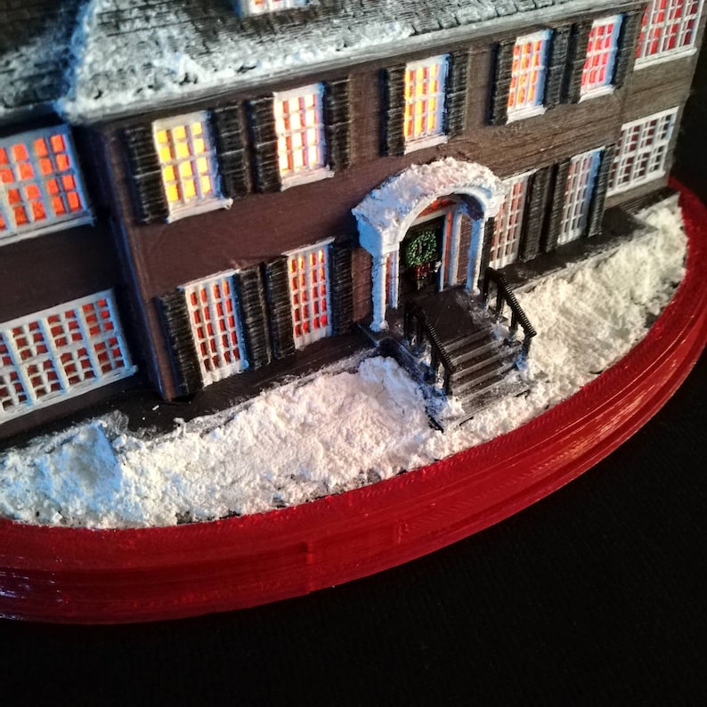 May include: A miniature model of a house with a red roof and white snow on the ground. The house has lit windows and a front porch with a wreath on the door. The house is sitting on a red base.
