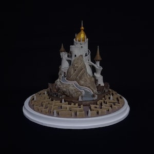May include: A detailed model of a castle with a gold dome, white towers, and brown accents. The castle sits atop a rocky mountain with a winding path, surrounded by a maze-like structure on a white base. The background is black.