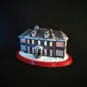 May include: A miniature replica of a two-story house with a snow-covered roof and lit windows. The house is sitting on a red oval base with a snowy landscape.