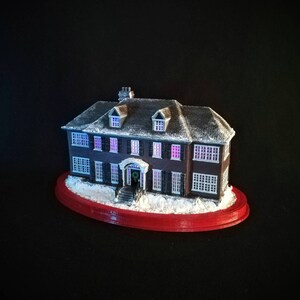 May include: A miniature replica of a two-story house with a snow-covered roof and a red base. The house has a front porch with a wreath on the door and windows with glowing purple lights.