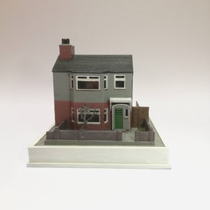 May include: A 3D printed model of a two-story house with a gray exterior, red brick accents, and a green door. The house has a small yard with a fence and a tree. The model is displayed on a white base.