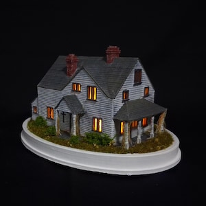 Op de afbeelding: Miniatuurmodel van een huis met twee verdiepingen, grijze buitenkant, donkergrijs dak en rode bakstenen schoorstenen. De ramen stralen warm licht uit. Het huis staat op een witte, ovale basis met groen.