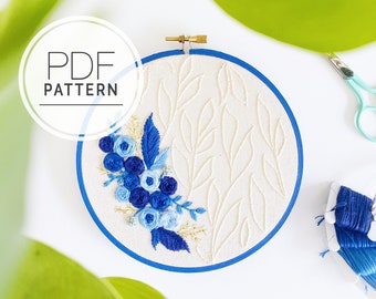Puede incluir: Un bastidor de bordado con un diseño floral azul y blanco. El diseño presenta flores y hojas azules, con el texto "PDF PATTERN" en un círculo blanco. El bastidor tiene un borde azul y está sobre un fondo blanco.