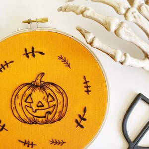 K&ouml;nnte beinhalten: Ein orangefarbener Stoff-Stickrahmen mit einem schwarzen K&uuml;rbis-Design. Der K&uuml;rbis ist von schwarzen, blattartigen Stickereien umgeben. Eine Skeletthand und eine schwarze Schere befinden sich im Hintergrund.
