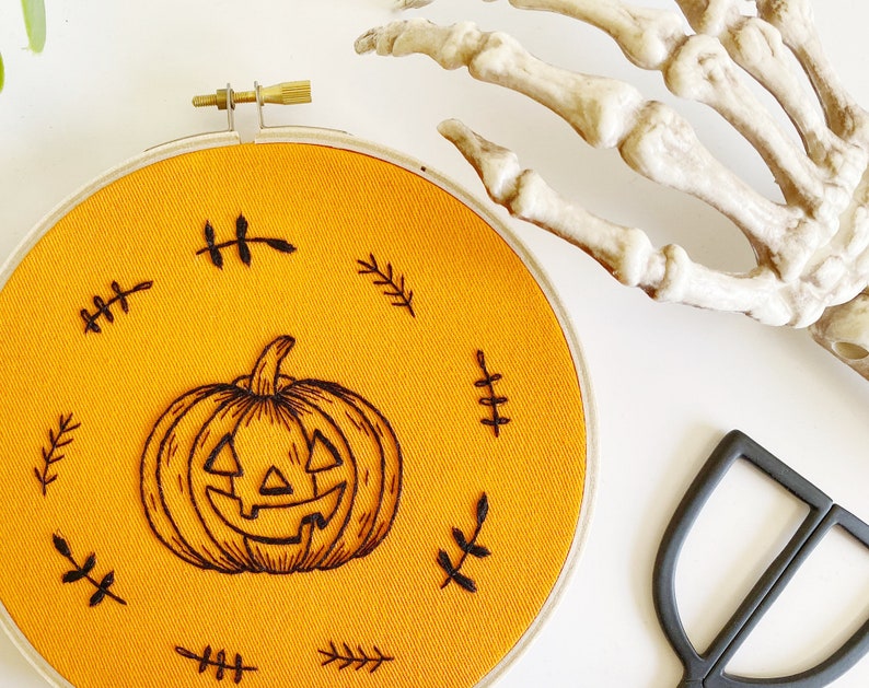 K&ouml;nnte beinhalten: Ein orangefarbener Stoff-Stickrahmen mit einem schwarzen K&uuml;rbis-Design. Der K&uuml;rbis ist von schwarzen, blattartigen Stickereien umgeben. Eine Skeletthand und eine schwarze Schere befinden sich im Hintergrund.