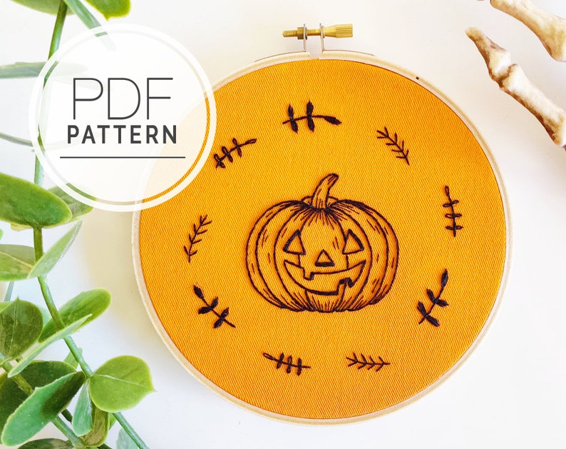 K&ouml;nnte beinhalten: Ein orangefarbener Stoff-Stickrahmen mit einem geschnitzten K&uuml;rbis-Design mit einem l&auml;chelnden Gesicht. Das Design ist von kleinen schwarzen, blattartigen Stichen umgeben. Der Text "PDF PATTERN" befindet sich in einem wei&szlig;en Kreis oben links.