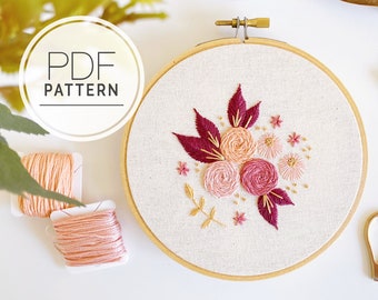 May include: A floral embroidery hoop with a cream-colored fabric background. The design features pink and burgundy flowers and leaves. The text "PDF PATTERN" is in a circle at the top left. Two spools of peach-colored thread are in the lower left.