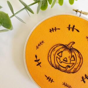 K&ouml;nnte beinhalten: Ein runder, orangefarbener Stoff-Stickrahmen mit einem geschnitzten K&uuml;rbis-Design. Der K&uuml;rbis ist von kleinen, schwarzen, blattartigen Akzenten umgeben. Der Stickrahmen ist mit einem Messingverschluss befestigt.