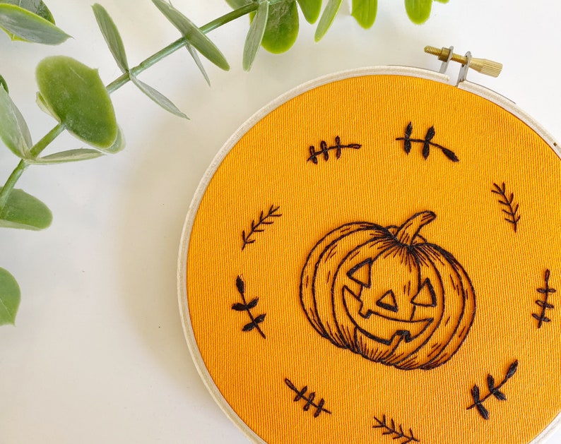 K&ouml;nnte beinhalten: Ein runder, orangefarbener Stoff-Stickrahmen mit einem geschnitzten K&uuml;rbis-Design. Der K&uuml;rbis ist von kleinen, schwarzen, blattartigen Akzenten umgeben. Der Stickrahmen ist mit einem Messingverschluss befestigt.
