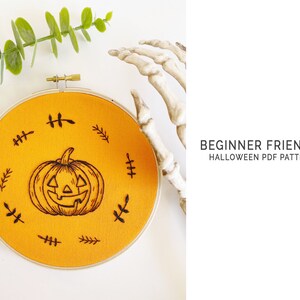 K&ouml;nnte beinhalten: Ein runder, orangefarbener Stoff-Stickrahmen mit einem schwarzen K&uuml;rbis-Design. Das Design ist von kleinen, schwarzen blattartigen Mustern umgeben. Der Text "BEGINNER FRIENDLY HALLOWEEN PDF PATTERN" ist sichtbar.