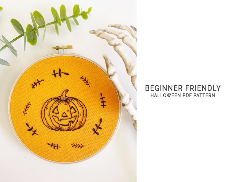 K&ouml;nnte beinhalten: Ein runder, orangefarbener Stoff-Stickrahmen mit einem schwarzen K&uuml;rbis-Design. Das Design ist von kleinen, schwarzen blattartigen Mustern umgeben. Der Text "BEGINNER FRIENDLY HALLOWEEN PDF PATTERN" ist sichtbar.