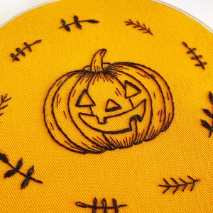 K&ouml;nnte beinhalten: Handgestickter Stickrahmen mit einem grinsenden K&uuml;rbis-Design in schwarzem Faden auf leuchtend orangefarbenem Stoff. Das Design ist von kleinen, gestickten Blattakzenten umgeben, passend zum Halloween-Thema.