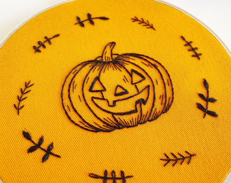 K&ouml;nnte beinhalten: Handgestickter Stickrahmen mit einem grinsenden K&uuml;rbis-Design in schwarzem Faden auf leuchtend orangefarbenem Stoff. Das Design ist von kleinen, gestickten Blattakzenten umgeben, passend zum Halloween-Thema.