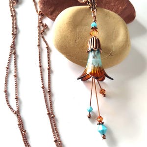 May include: A copper-colored necklace with a pendant shaped like a stylized flower. The flower pendant has blue and orange enamel, with turquoise and amber-colored beads. The necklace is displayed on a white surface.