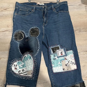 May include: Blue denim jeans with Mickey Mouse patches and embroidery. The jeans have a heart-shaped patch on the left leg and a rectangular patch on the right leg. The cuffs are decorated with Mickey Mouse fabric and camouflage fabric.