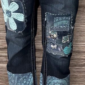 Può includere: Jeans in denim blu scuro con dettagli di patchwork, tra cui un fiore all'uncinetto e tessuto paisley. I jeans hanno un aspetto consumato e sono risvoltati in fondo. La persona indossa sandali in denim con un'etichetta marrone che dice "Silver".