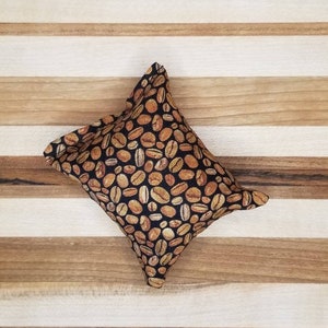 May include: A small, diamond-shaped fabric item with a black background covered in tan coffee bean illustrations. The item is resting on a light and dark wood striped surface.