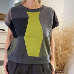 May include: A short-sleeved top featuring a colour-blocked design with various shades of grey, dark charcoal, and olive green. A prominent, vertical, lime green geometric shape resembling a stylised vase or bottle is centred on the front.