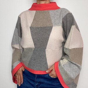 May include: A long-sleeved pullover jumper featuring a patchwork design of geometric shapes in shades of grey, beige, and cream. The garment has a bright coral-coloured mock neck collar and matching wide cuffs at the ends of the sleeves.