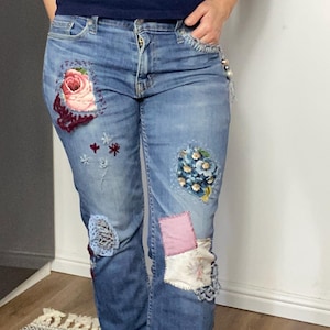 May include: Light blue denim jeans with floral patches. Patches include a large pink rose, blue floral arrangements, and other fabric accents. The jeans have a classic five-pocket design and a button and zip closure.