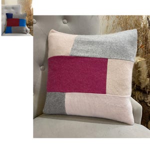 May include: A square throw pillow featuring a color-blocked design with rectangular panels in shades of soft pink, light gray, and a deep magenta. The fabric has a textured, knit-like appearance with visible seams connecting the distinct color sections.