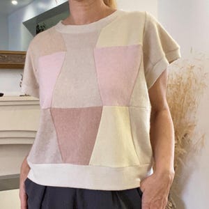 Könnte beinhalten: Ein kurzärmeliges Oberteil mit einem geometrischen Patchwork-Design in sanften Pastelltönen wie Creme, Blassrosa, Hellgelb und gedämpftem Beige. Das Kleidungsstück hat einen Rundhalsausschnitt, umgeschlagene Ärmel und einen geraden Saum mit cremefarbenem Bund.