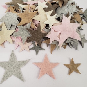 May include: A collection of die-cut felt stars in various sizes and colors, including shades of light pink, cream, tan, dark brown, and light gray. The stars are arranged in a large pile with three individual stars placed in the foreground.