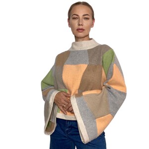 May include: A long-sleeve jumper featuring a patchwork design with large geometric panels in shades of light green, peach, tan, and grey. The garment has a high mock neck collar and wide, flared sleeves with cream-coloured trim at the cuffs.