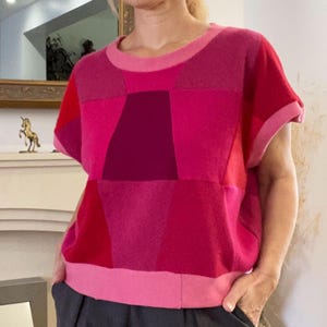 May include: A short-sleeved knit top featuring a geometric patchwork design in various shades of pink, magenta, and red. The garment has a round neckline and a wide, solid light pink hem band at the waist, with cuffed sleeves in a matching light pink.