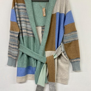 May include: A patchwork cardigan in shades of blue, brown, green, and beige. The long-sleeved sweater has a shawl collar and a matching belt. The word "SLOW" is printed on a tag hanging from the collar.